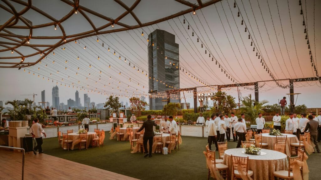 Elegant rooftop event with tables and lights set against Mumbai skyline at sunset, India.